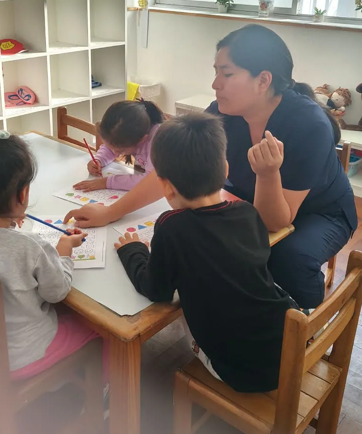 La terapeuta acompaña a tres niños trabajando con fichas en la mesa y le da retroalimentación a uno en el momento